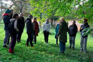 Rosemary Lee is stood in an orchard talking to a group of 11 people stood around her. wearing glasses, a green parka coat, blue jeans and a yellow hat loosely on her head.