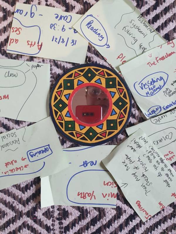 a sheet of white paper, in which Lateisha wrote the workshop plan on, is cut up and spread around a small mirror. The mirror has a geometric patterned frame in yellow, green, red and blue. In the mirror is Lateisha's face and phone taking a selfie