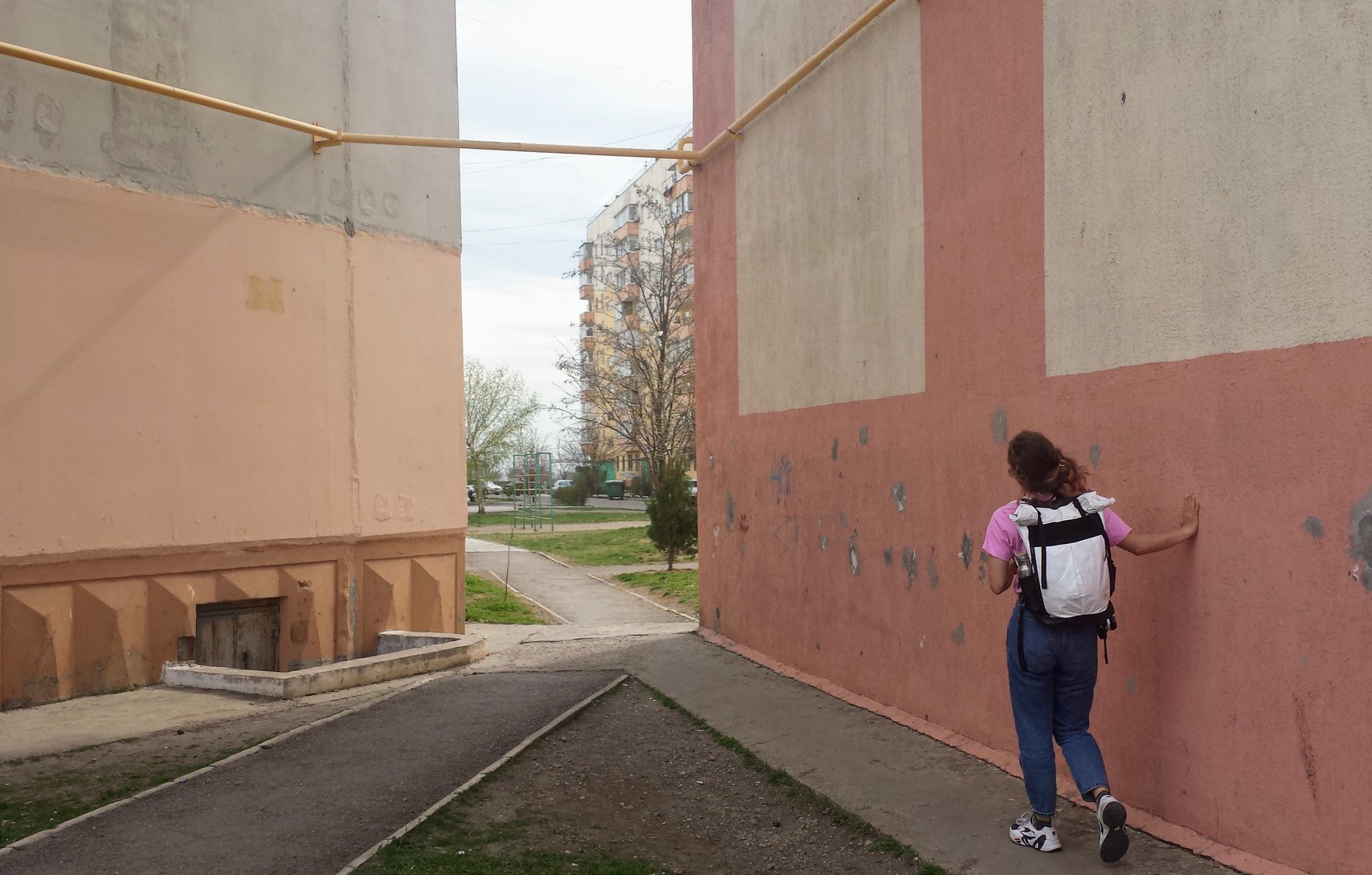 A shot of a person walking away through a path between two buildings. The person is wearing a pink t-shirt, dark blue jeans and a black and white backpack, they have their right hand tracing alongside the dark rose wall beside them.