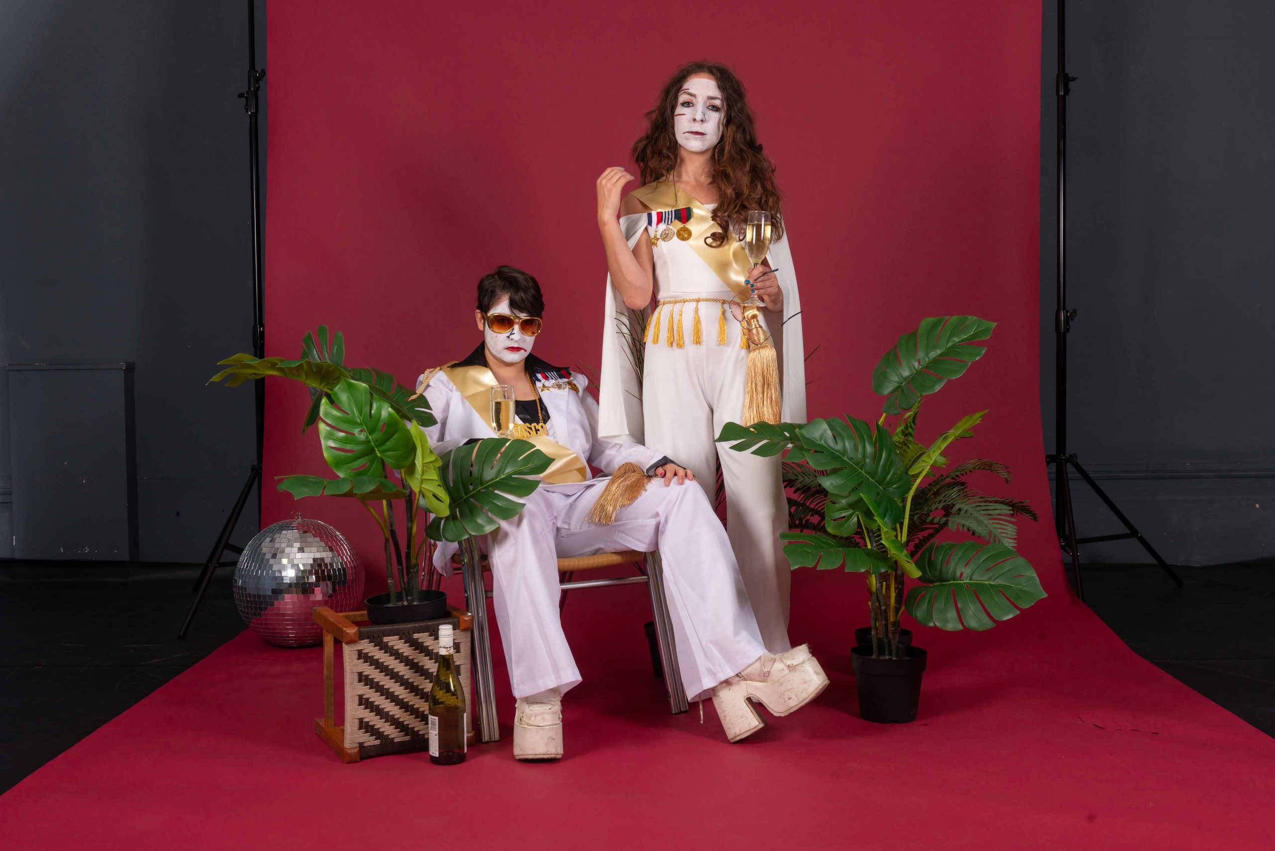A photo of two people, both wearing white outfits with gold satin sashes and a lapel with multiple medals. Their faces are painted white, as they both hold filled champagne flutes. One person is sitting, wearing gold-toned sunglasses with one leg lengthened forward, wearing white platform heels. The other person is standing above them with wavy brown hair, staring straight into the camera. They are standing on a rolled maroon-coloured backdrop with two potted plants and an empty bottle of wine by their feet, with a disco ball on the floor. 