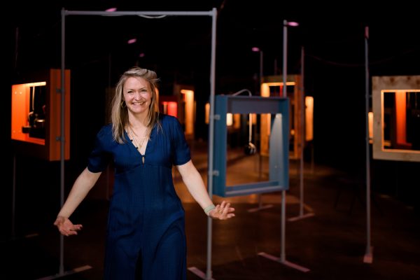 A person stood smiling with blond hair wearing a midnight-blue dress. In the background, out of focus is an installation of wooden frames, all varying in size, colour and height, held together by metal poles.