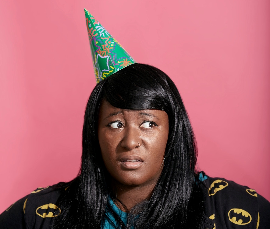 A headshot of a person wearing a green colourful party hat against a pink backdrop as they side-eye. The person is wearing a black cardigan with Batman-insignia, and they have long black hair. 