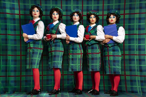 A photo of five people stood in unison in front a green tartan backdrop, they are all wearing identical tartan tuxedos with skirts, white shirts, red tights and black shiny dress shoes. The faces are all painted with identical blue eyeshadow, red squiggly eyebrows, and red lipstick. Two of them are holding red mugs and three are holding blue folders.