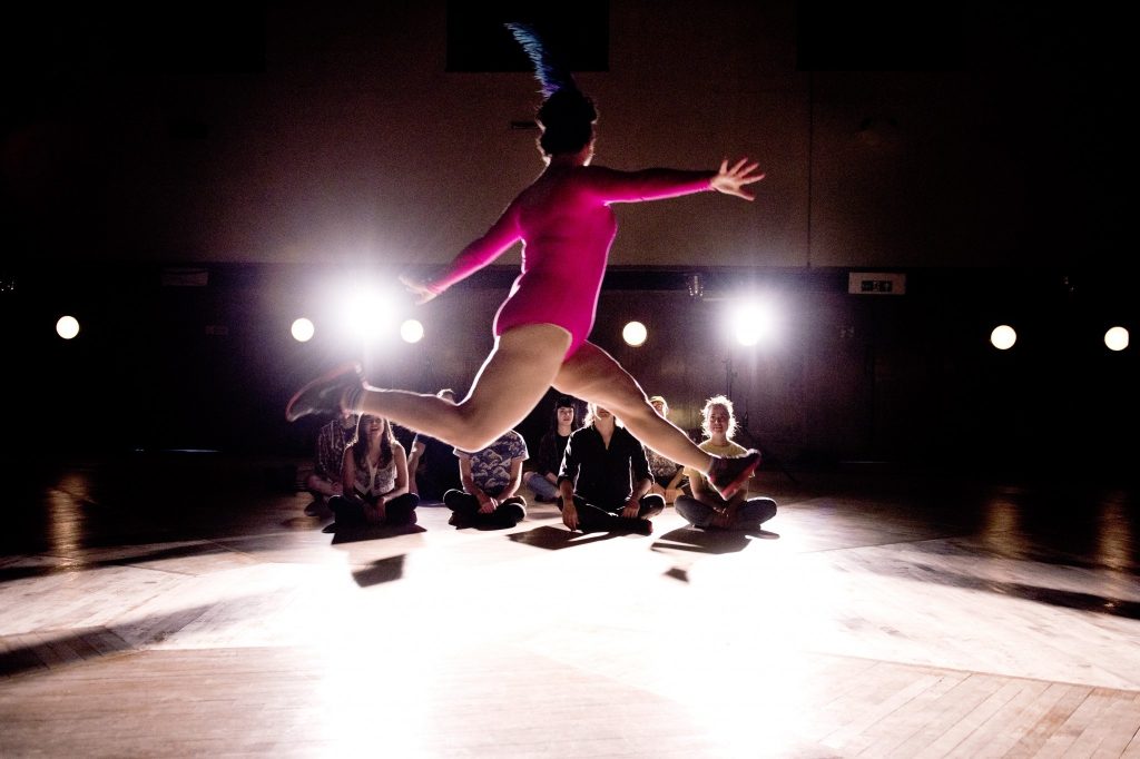 A shot of a person dancing in the air, as they leap across the room with both arms and legs outstretched. They are wearing a hot pink leotard with a blue feather on their head. Stage lights are gleaming behind them as a small audience sits on the floor watching them. 
