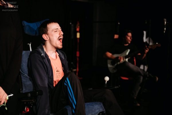 A person wearing an orange t-shirt, black hoodie and joggers in their wheelchair with their mouth wide open, performs against a dark background. Also, in the frame there is an out of focused person playing the guitar. 