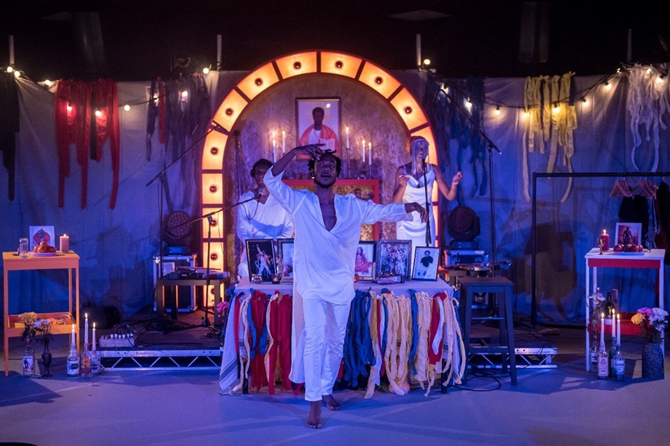 A person performing on a stage wearing a white kaftan top with white trousers, they are mid pose with arms outstretched. There is a band of two people behind them, both also wearing white as they sing into microphones. The performers are in front of an altar, adorned with colourful fabric streamers, behind them gleaming festoon lights and on the floor are candles and flowers.
