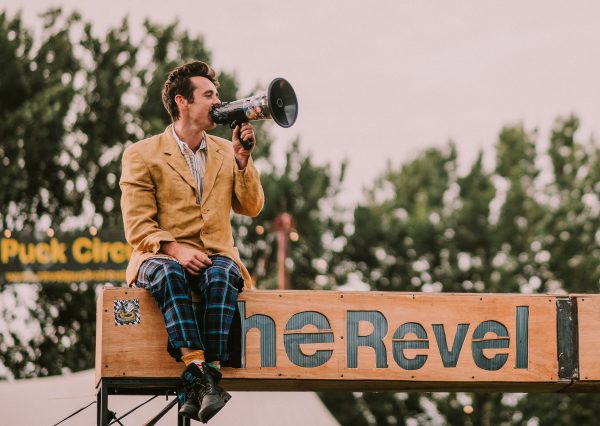 A person sat high on top of a wooden sign speaking into a megaphone. The sign reads 'The revel’… The person is wearing a beige suit and blue tartan trousers with combat shoes, in the background are trees and empty skies.