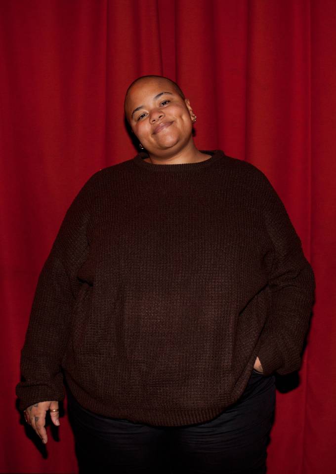 Nene is pictured here smiling against a red background. They are person with a light brown complexion and they are wearing, a brown woolly jumper.
