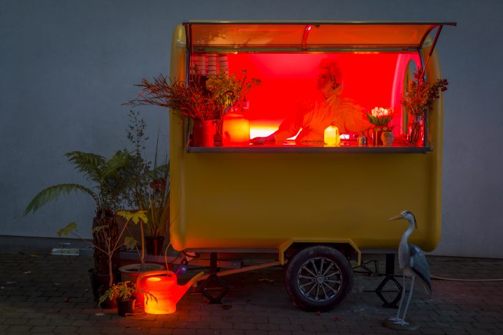 Jennie Moran, stood inside a yellow food cart with a red neon glow inside. In front of the cart is an illuminated red watering can
