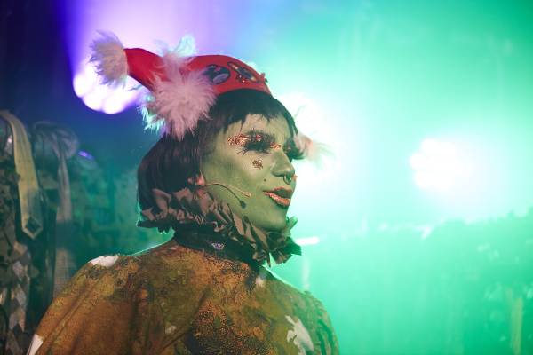A photo of Oozing Gloop performing, Her face is painted green with gold fragmented foil makeup on her eyelid and cheeks, with red lipstick. She’s wearing a white and brown long-sleeved cow pattern dress and a red pirate hat that has white feathers on either side. Blue and green stage lights are gleaming behind her