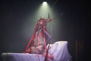 A photo of Bonnie Bakeneko performing. They are knelt on a white medical bed with their head tilted back against a dark background with blood stains all over them and the bed. They are wearing a cream undergarment and a red horn headpiece with long red strings and jewels hanging off it.