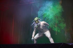 A photo of Shrek666 performing. He is a green creature with two horns and a long light purple beard. He is leant forward with one arm on his back and one arm hanging. The set contains soft green and red lights, with green smoke appearing behind him