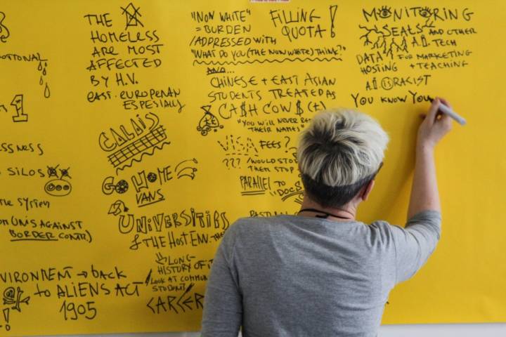 A person with short blonde hair and a grey top writes on a big piece of yellow paper