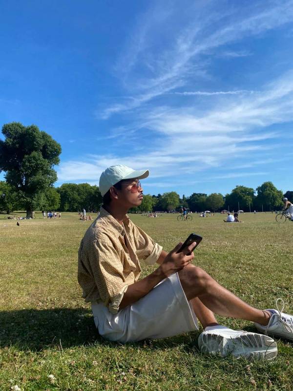 Benji - a man with short dark hair sits on the grass on a beautifully sunny day, wearing a mint green cap, long white shorts, a beige patterned shirt, and a pair of sunglasses. He is framed in profile, sitting cross-legged and facing to the right.