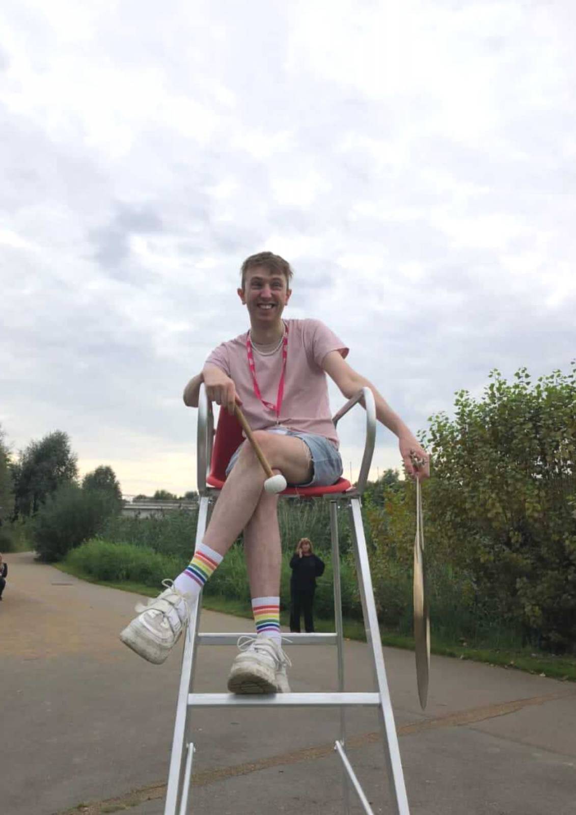 Aidan, who wears a pink t-shirt, denim shorts, rainbow coloured socks and white trainers, sits on a tall umpire's chair and laughs.