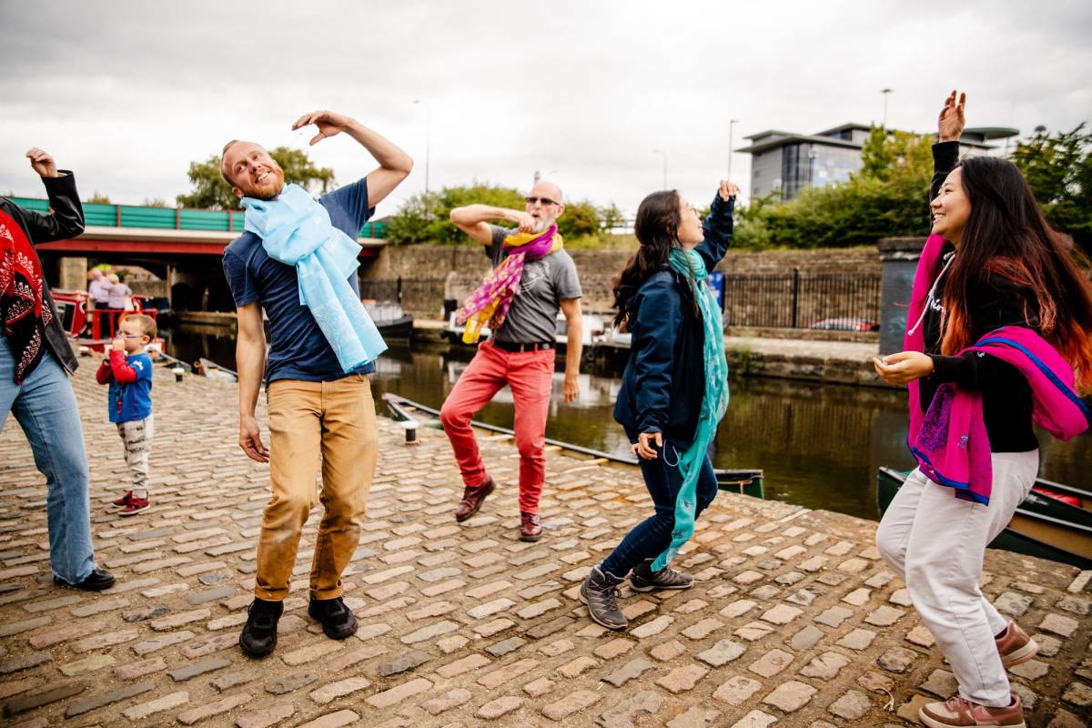 A group of people practice Bhangra dancing together, wearing colourful scarves, by the side of the canal.