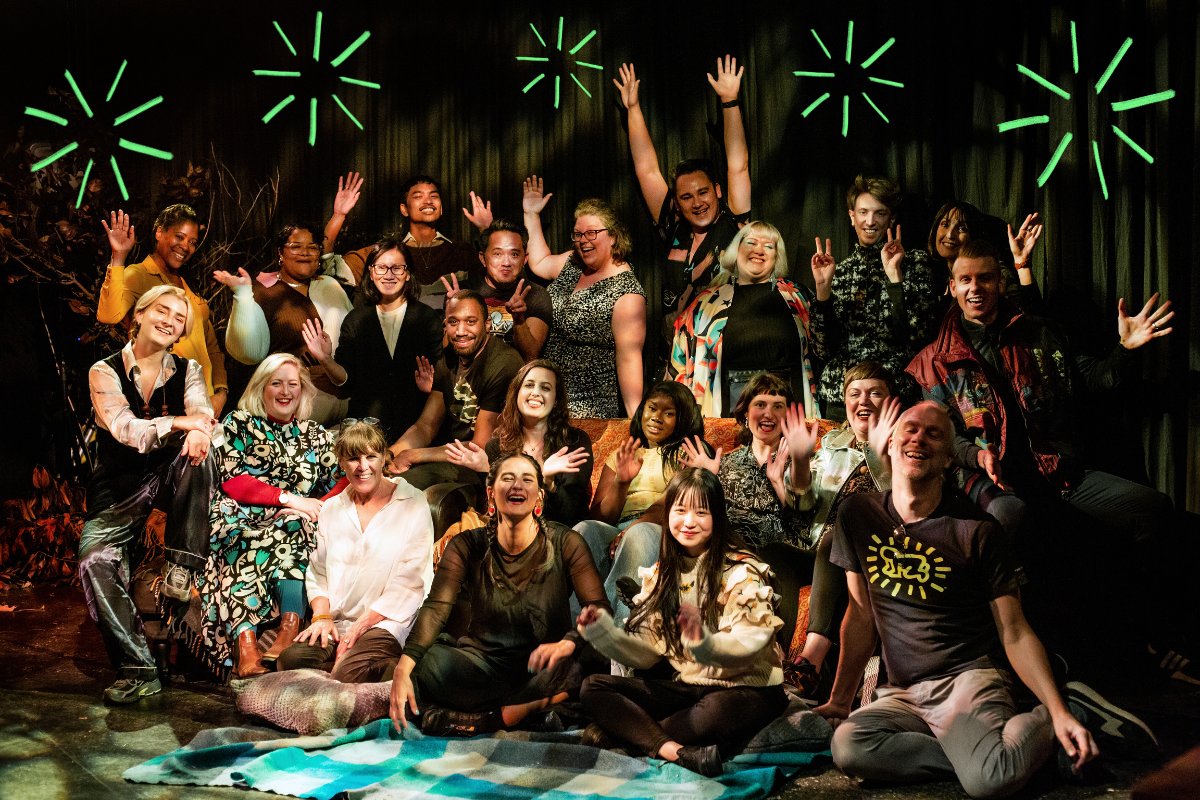 The Artsadmin team gather for a group photo in the theatre, against a dark curtain and surrounded by foliage and dappled autumn lighting. Some sit in the foreground on a blue checked rug, some sit on chairs and sofas behind, and some stand at the back. Everyone is wearing their party clothes and smiling, and the photo is illustrated with neon green fireworks.