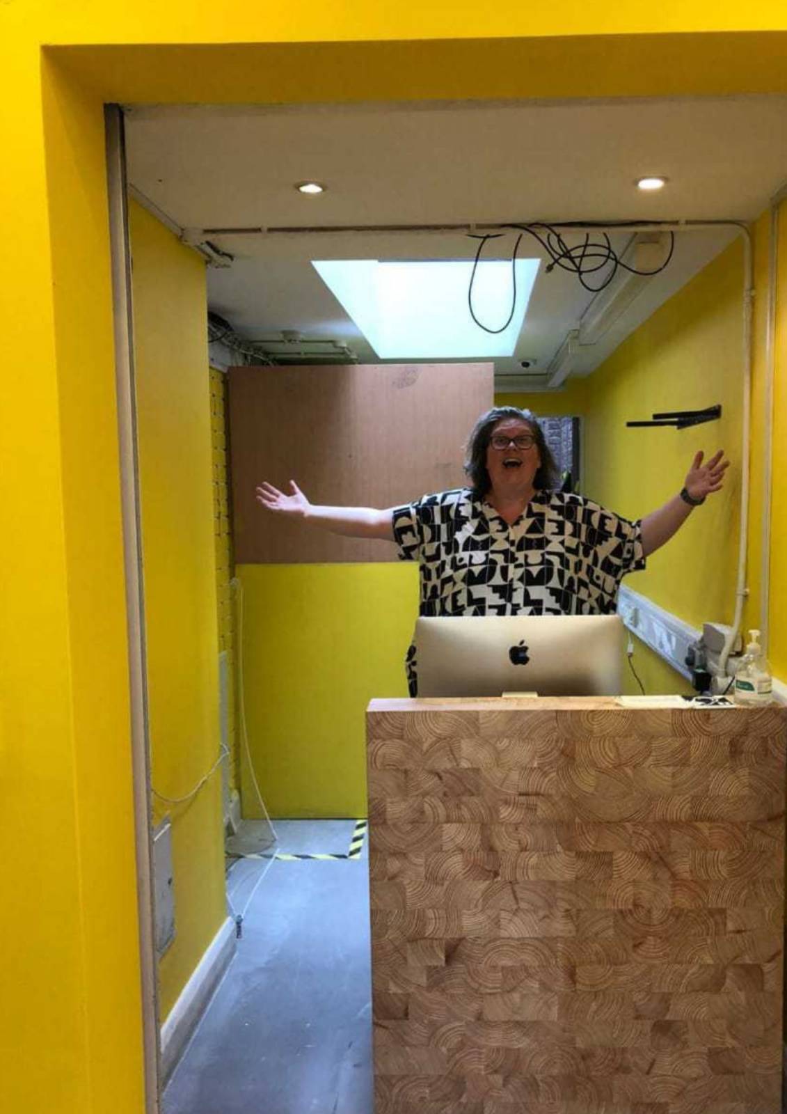 A woman wearing a black and white patterned top raises both arms in a gesture of welcome and excitement, as she stands behind a reception desk at Toynbee Studios. The walls around have been newly painted bright yellow. 
