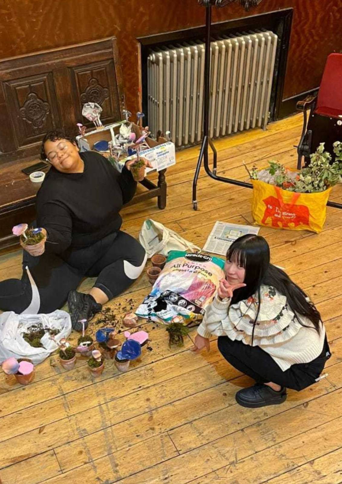 Two young people - Nene and Zoey, sit or crouch on a wooden floor in the middle of a crafts session at Toynbee Studios. They are both smiling for the camera, whilst they make mushroom sculptures out of newspapers.