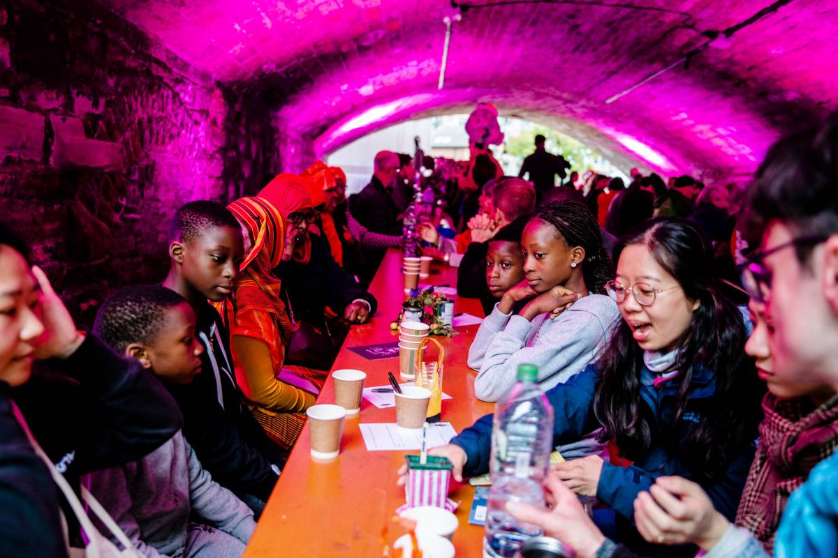 A large group of people enjoy a meal together inside a tunnel lit with hot pink lights.