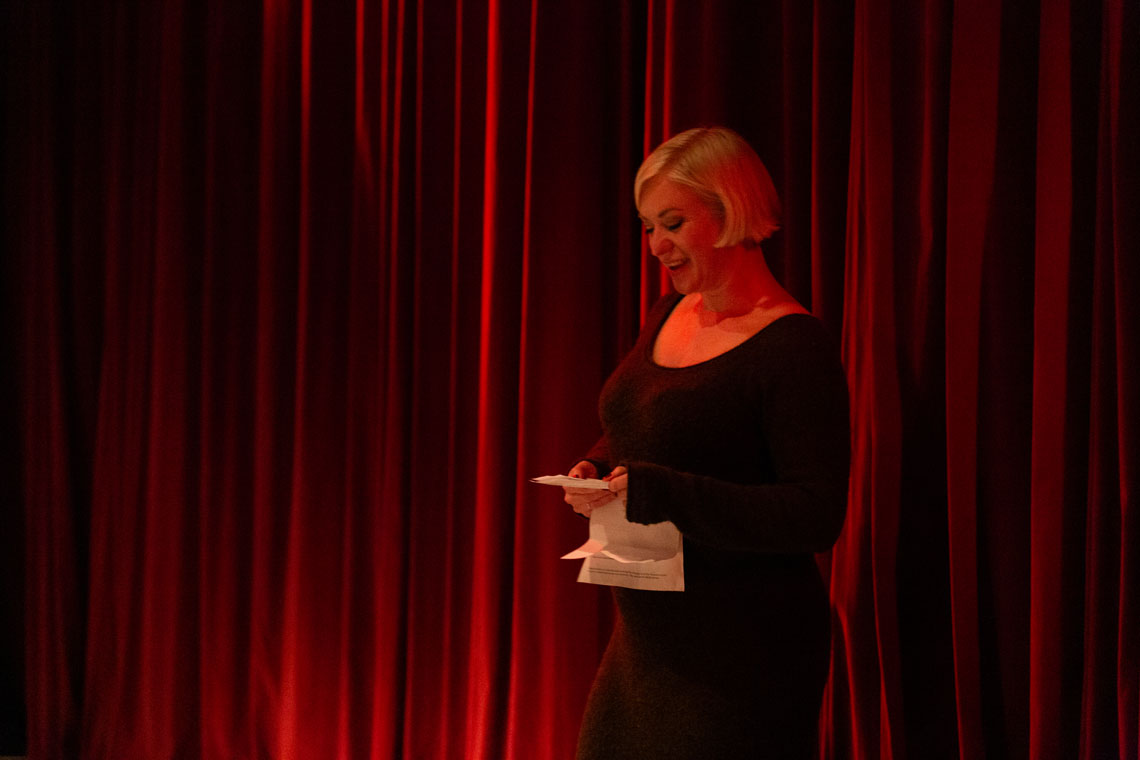 Jennie Moran, a white woman with short blonde hair, gives a toast. She stands against a red velvet curtain