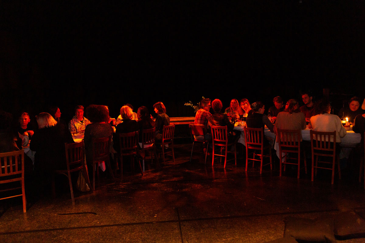 Two tables converge, dimly lit, as the guests sit down to eat