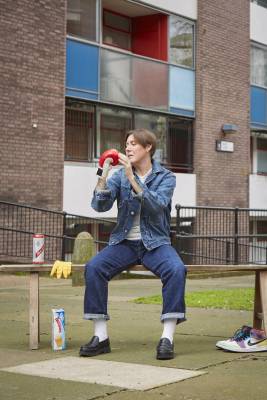 An image of Tink Flaherty sitting on a wooden bench in a council estate. They wear double denim, white socks and black loafers, and have a boxing glove on their right hand, which they are gazing at and caressing with their left hand. Their brown hair is short, and falls slightly over their right eye. Around the bench are a pair of trainers, a glove, and a packet of Twinkies