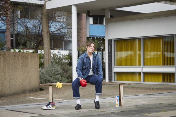 An image of Tink Flaherty sitting on a wooden bench in a council estate, facing the camera. They wear double denim, white socks and black loafers, and have a boxing glove on their right hand. Their brown hair is short, and falls slightly over their right eye. Around the bench are a pair of trainers, a glove, and a packet of Twinkies