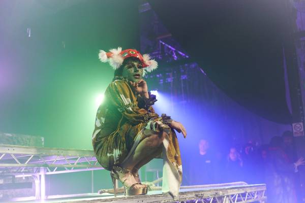 A photo of Oozing Gloop performing on a stage, Her face is painted green with gold fragmented foil makeup on her eyelid and cheeks, with red lipstick. She’s wearing a white and brown long-sleeved cow pattern dress, a red pirate hat that has white feathers on either side, and gold platform heels. She is crouched down looking into the audience with one hand resting on her legs and one hand against her chin. Blue and green stage lights are gleaming behind her as a small crowd is stood beside the stage watching her.