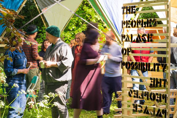 An image of brightly dressed people in a geodesic dome structure outdoors, milling around and talking. A wooden sign on the right says 'The People's Palace of Possibility'