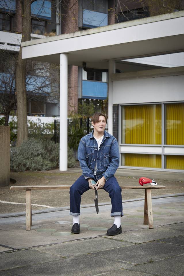 An image of Tink Flaherty sitting on a wooden bench in a council estate. They wear double denim, white socks and black loafers, and hold a knife in their hands, blade downwards, as they gaze into the camera