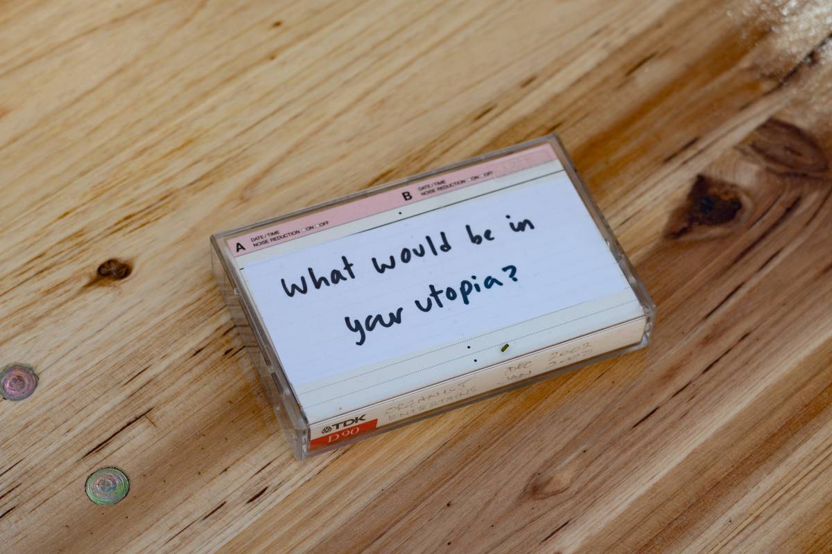 A photo of a cassette tape placed on a wooden table. On the tape is written 'What would be in your utopia?'