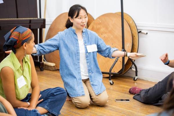 A photo of Haeweon Yi kneeling on a wooden floor with her arms outstretched, delivering a workshop to people sitting cross-legged on the floor