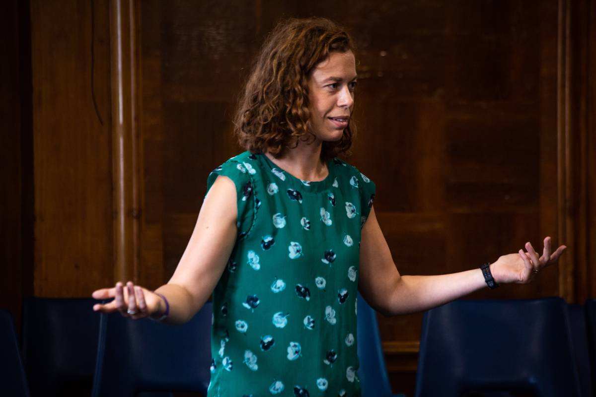 A photo of Katy Rubin, in the middle of delivering a Legislative Theatre workshop. She stands in a dark wooden room, making a bold questioning gesture