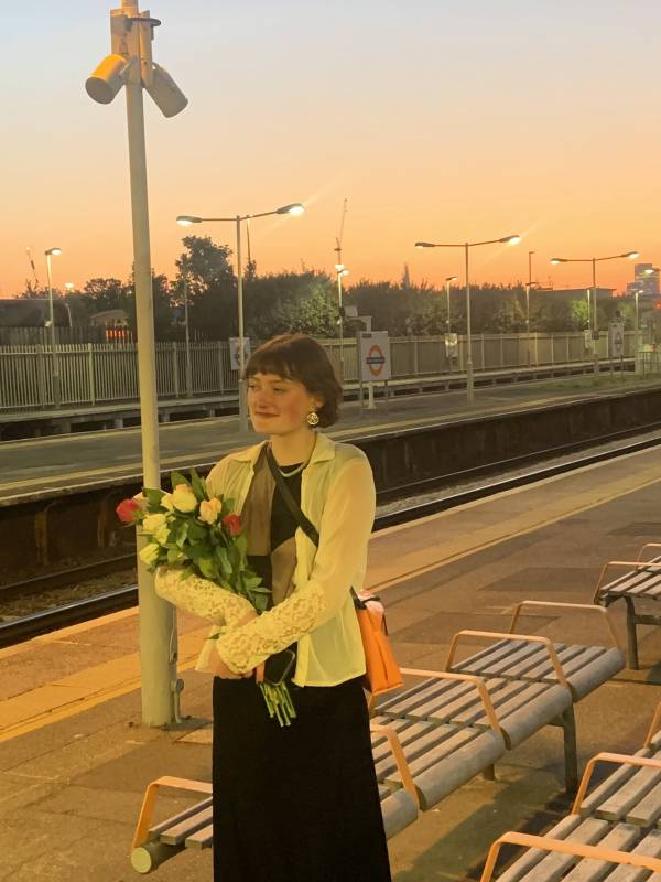 Daisy is standing on a platform at dusk smiling whilst holding a bouquet of flowers.