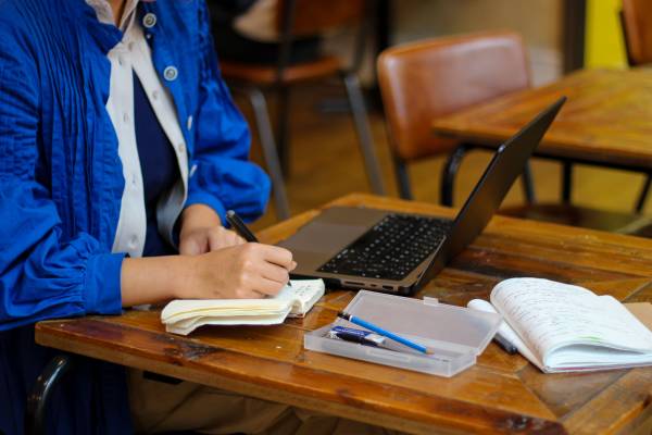 An image of a person from the neck down wearing a blue jacket and white top. They are at a desk writing in a notebook surrounded by an open laptop, a pencil case and another notebook