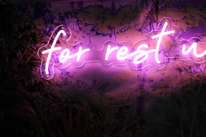 a closeup of a glowing pink neon sign, reading ‘for rest’ in a handwritten style font. The glow of the light is illuminating the plant printed wallpaper behind it.