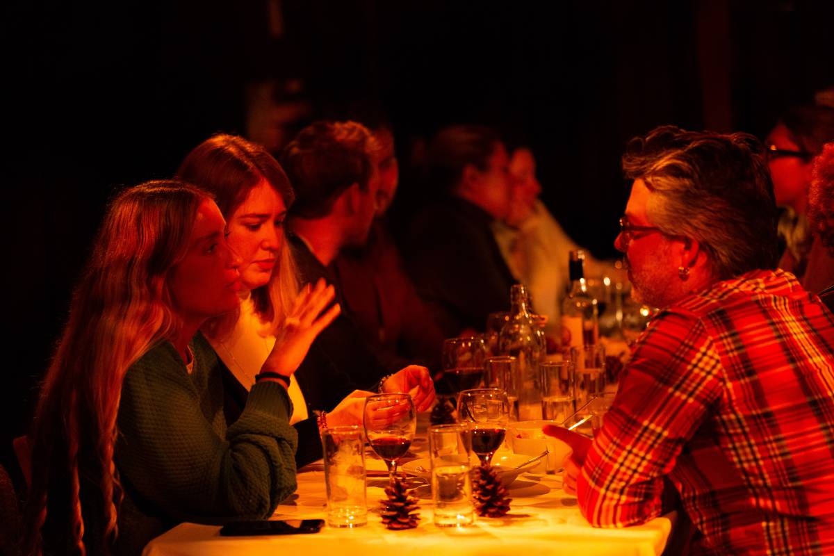 A photo of a warmly-lit yet dark table covered with food and drink, and people talking to one another