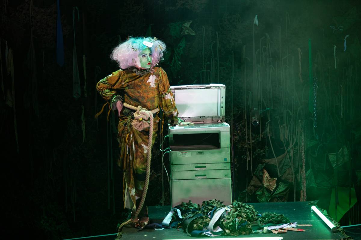 An image of the drag creature Oozing Gloop, who wears a swampy green costume and stands on a hazy, mulchy stage in front of an old printer
