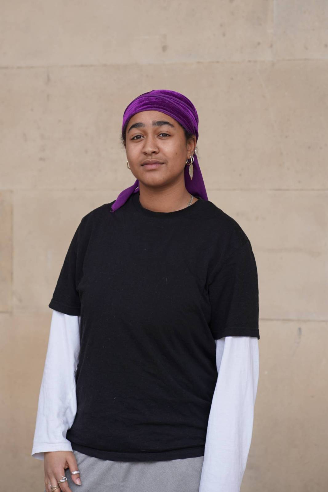 An image of Seyi standing in front of a beige stone background. They are wearing a purple head scarf tied at the back and a black tshirt with a white long-sleeve underneath.