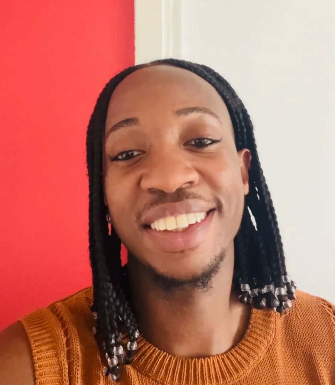 a selfie of An0maly smiling against a red and white background. He has his hair in shoulder length braids and wearing an orange sweater vest.