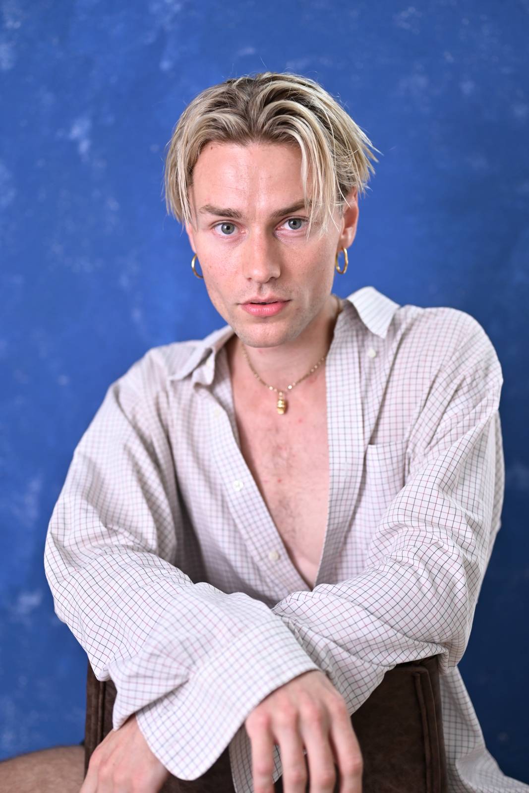 An image of Fraser from Raze collective with a blue background. They have blonde straight hair in a middle parting and are wearinga white shirt, unbuttoned down to their chest. 