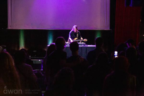 An image of a person standing on a stage lit with purple light. They are using a DJ deck whilist holding a guitar. There is a large crowd watching them in the foreground.