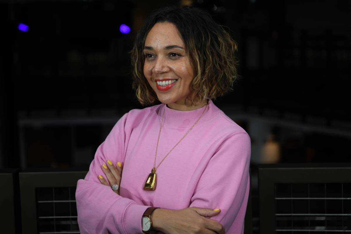 A photo of Raidene, a brown-skinned female, aged early forties, with wavy brown shoulder-length hair, wearing a pink jumper, looking away from the camera, smiling.