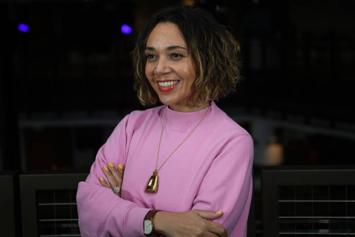 A photo of Raidene, a brown-skinned female, aged early forties, with wavy brown shoulder-length hair, wearing a pink jumper, looking away from the camera, smiling
