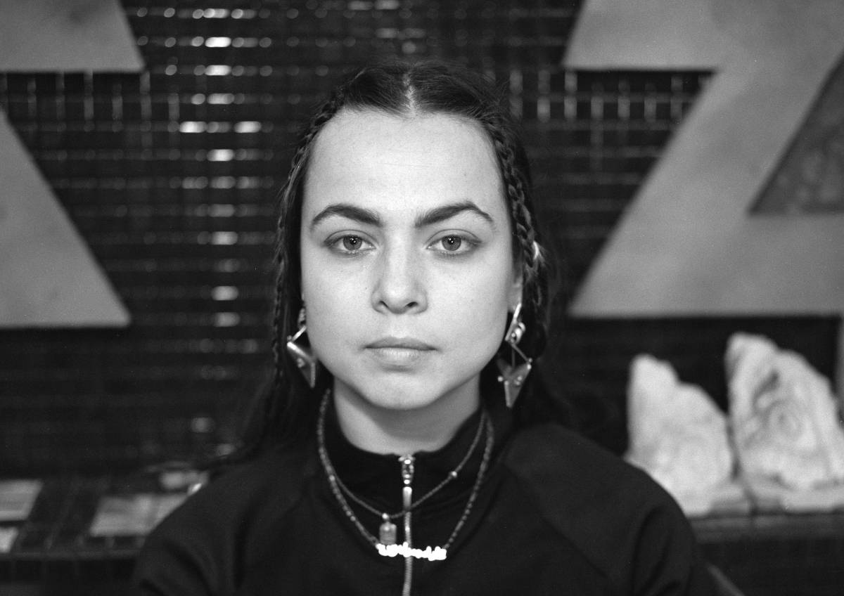 Black and white image of Alia Hamaoui in front of an artwork with a tiled surface and plaster casts. Alia is a cis gender woman with long brown hair, wearing a dark zip up top and chunky silver earrings and necklaces. She is looking directly at the camera. 