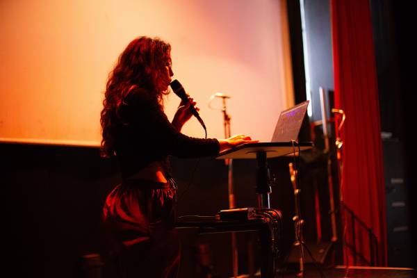 A low lit profile photo of a performer on stage holding a microphone in one hand and the other hand working a laptop. There is a large screen in the background.