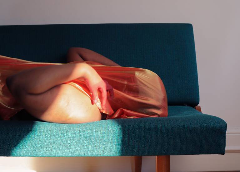 Bilan laying down on their back in the nude,on a dark green sofa with their arms covering their chest. Wearing an orange/golden iridescent scarf hiding their face.