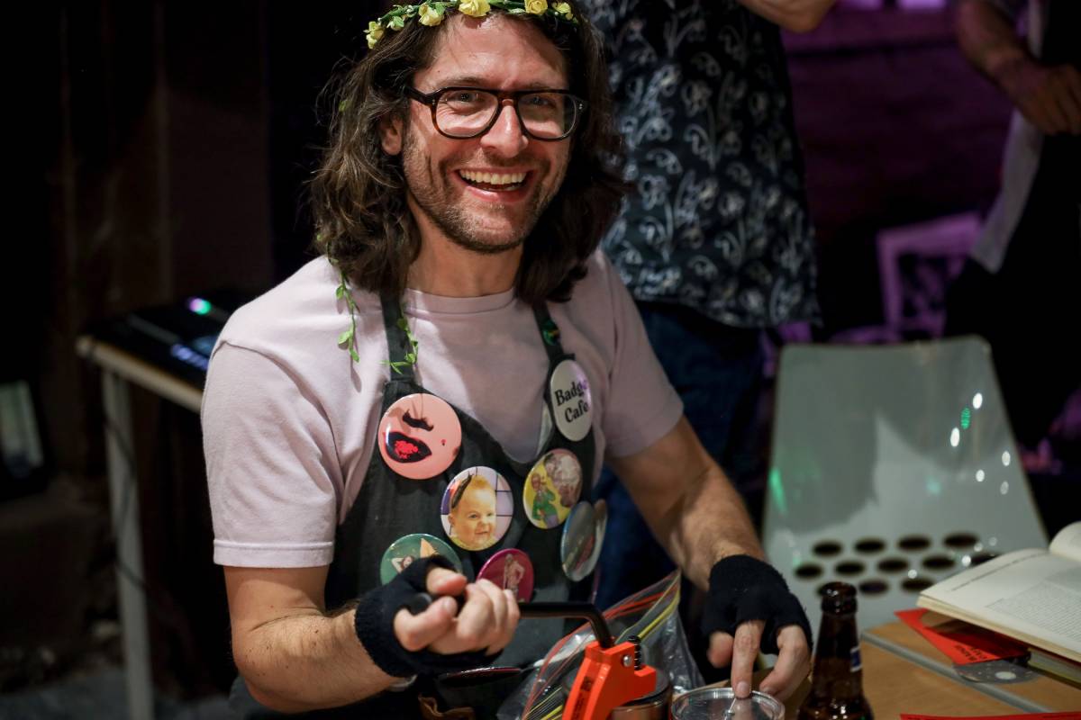 Badge Cafe host Ben Walters sits at his badgemaking station smiling at the camera. He has glasses and shoulder-length brown hair with a garland of yellow flowers in it. Ben is wearing an apron covered in colourful collaged badges over a lavender t-shirt and fingerless gloves.