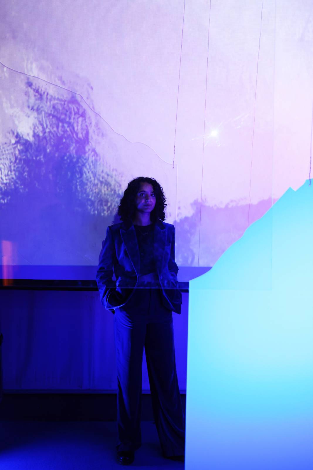 Jessica has short brown curly hair, wears a blue suit jacket and black top underneath. Her face and body are obscured by a purple projected image. Photo by Sara Benabdallah.